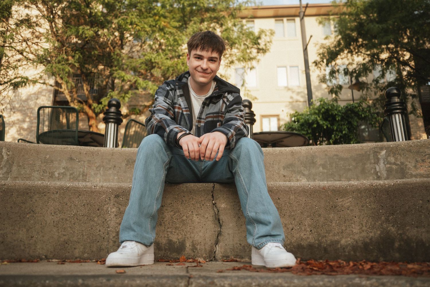 Person in casual attire sits relaxed on concrete steps with autumn foliage and buildings visible behind.