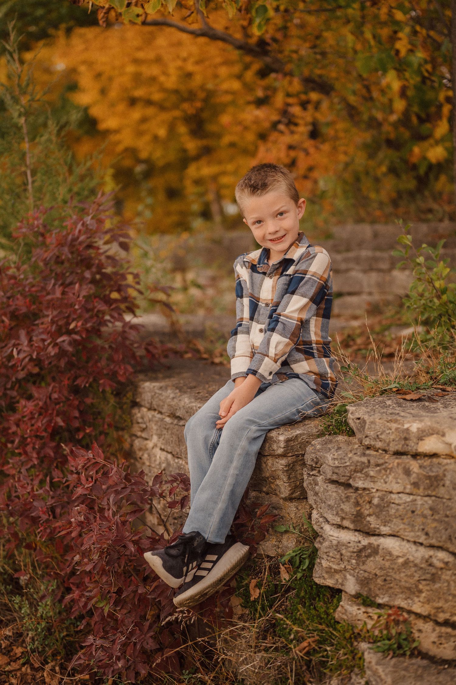 Child sitting on rocky ledge surrounded by vibrant autumn foliage in outdoor portrait session.