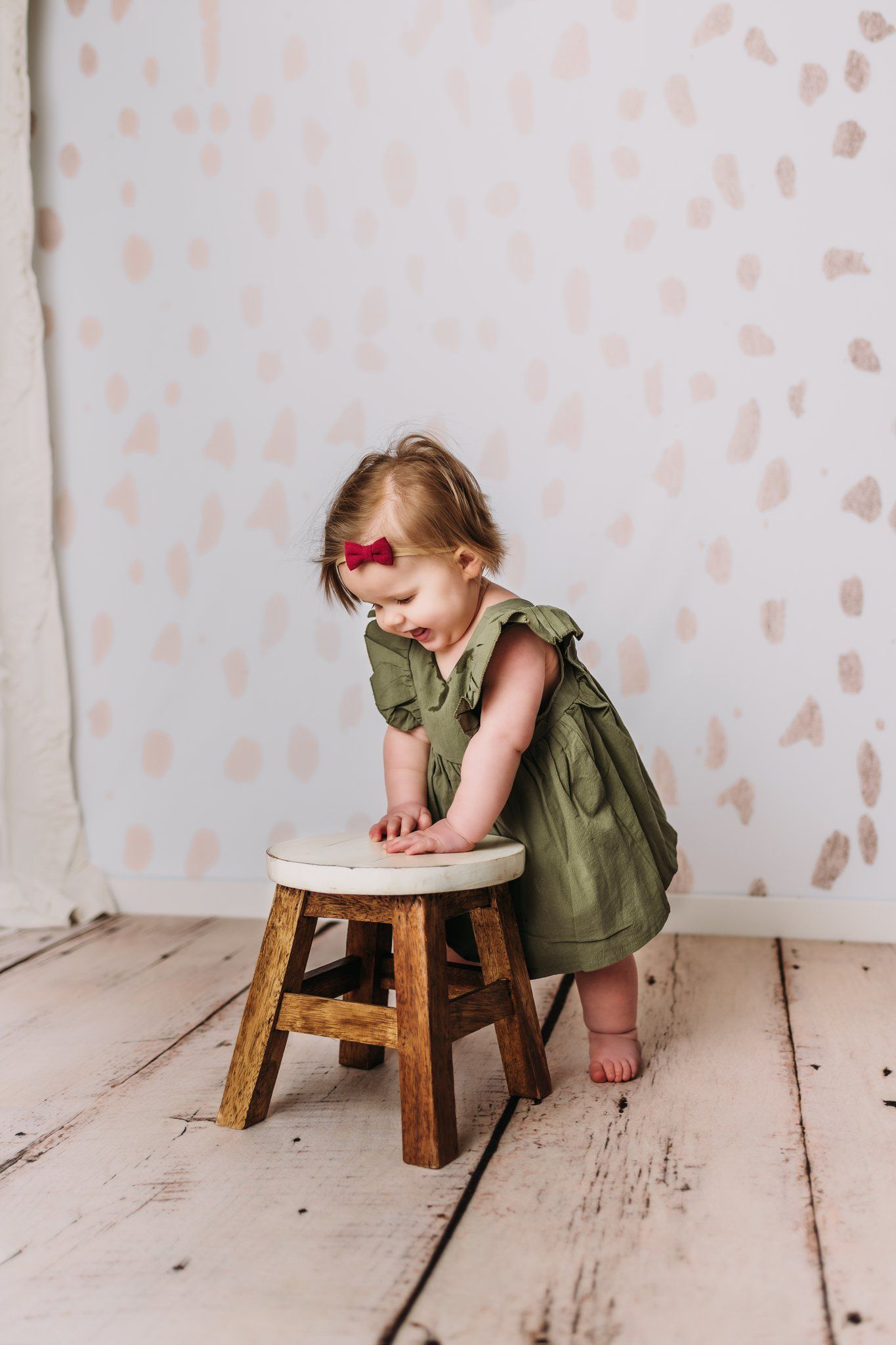 A small figure in an olive green dress leans over a wooden stool against a white backdrop with delicate pattern details.