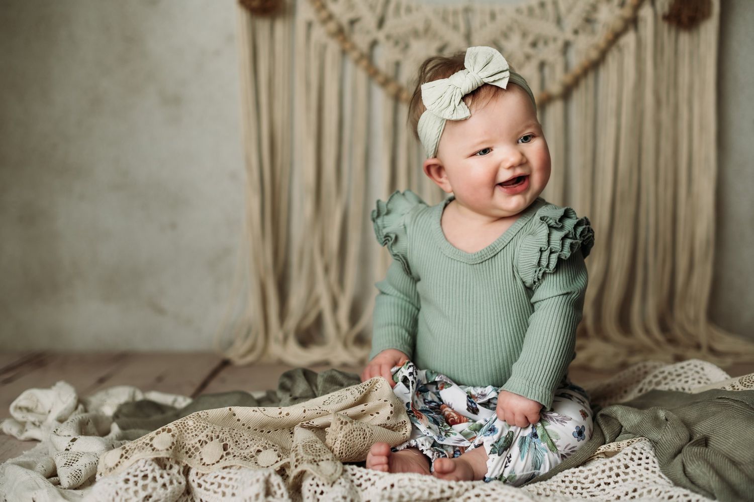 Happy baby in mint outfit and bow poses against boho macrame wall decor.