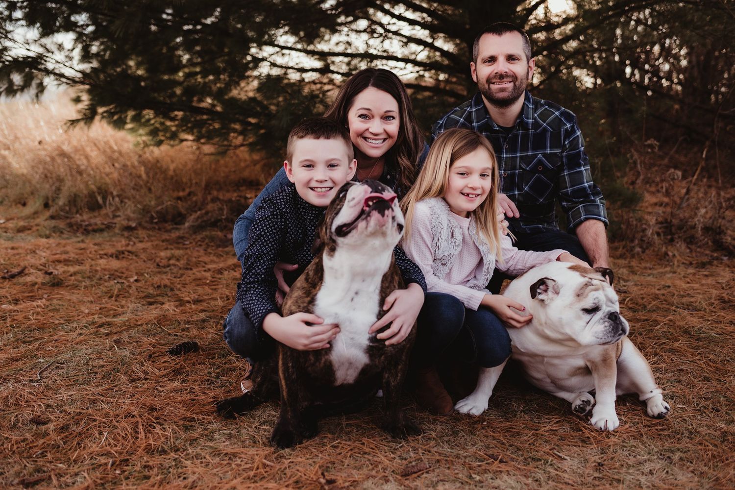 Family portrait outdoors with two white dogs surrounded by autumn foliage.