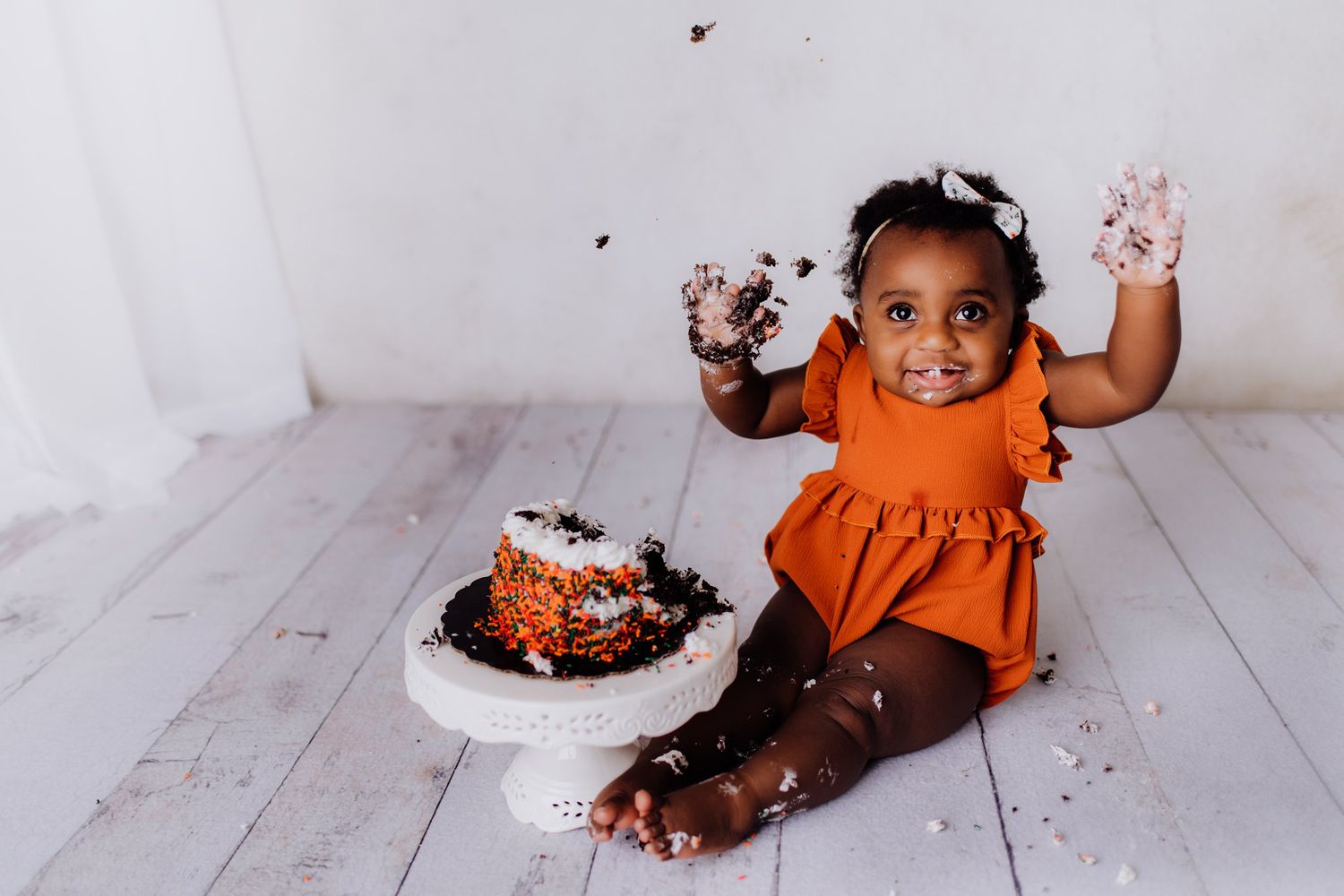 Baby in orange dress with hands raised during cake smash photo session on white wooden floor.