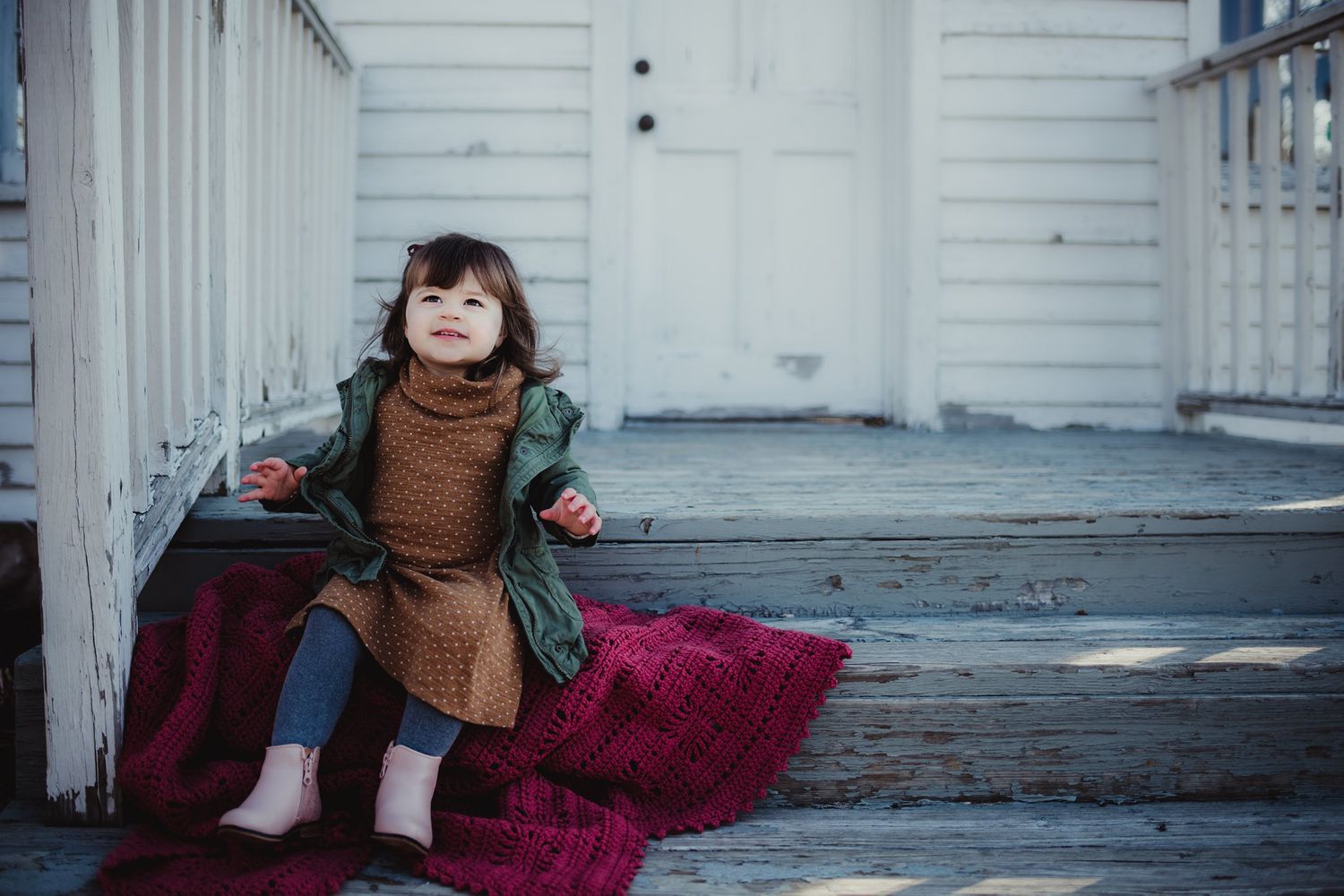 A toddler sits on white porch steps with a red blanket wearing a green jacket.