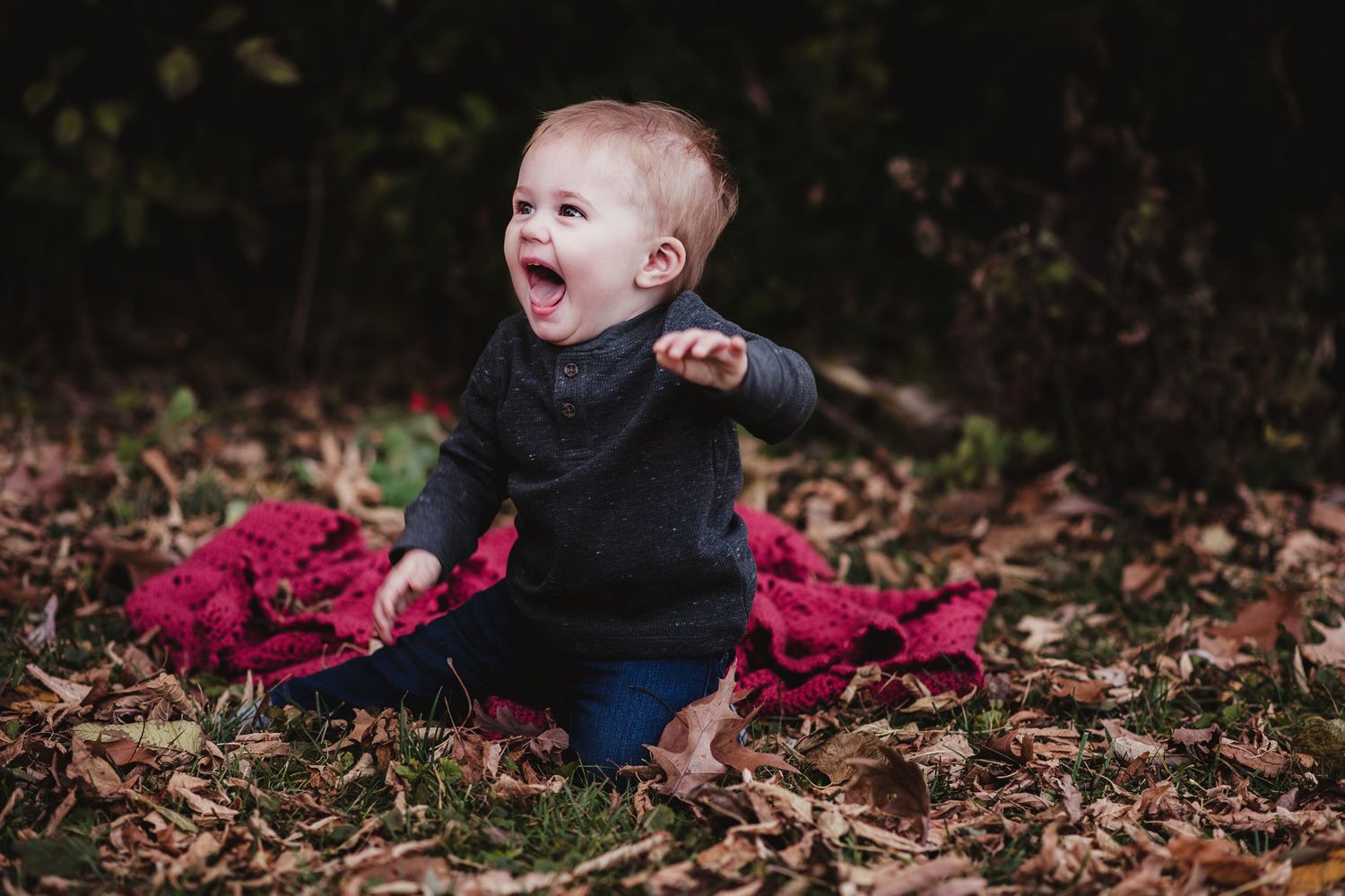 Small infant sits among fall leaves wearing dark clothing with bright pink accents.