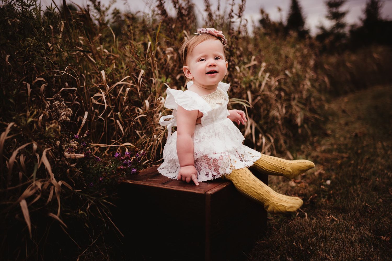 Baby in white dress and yellow socks sitting on rustic bench among tall grasses.
