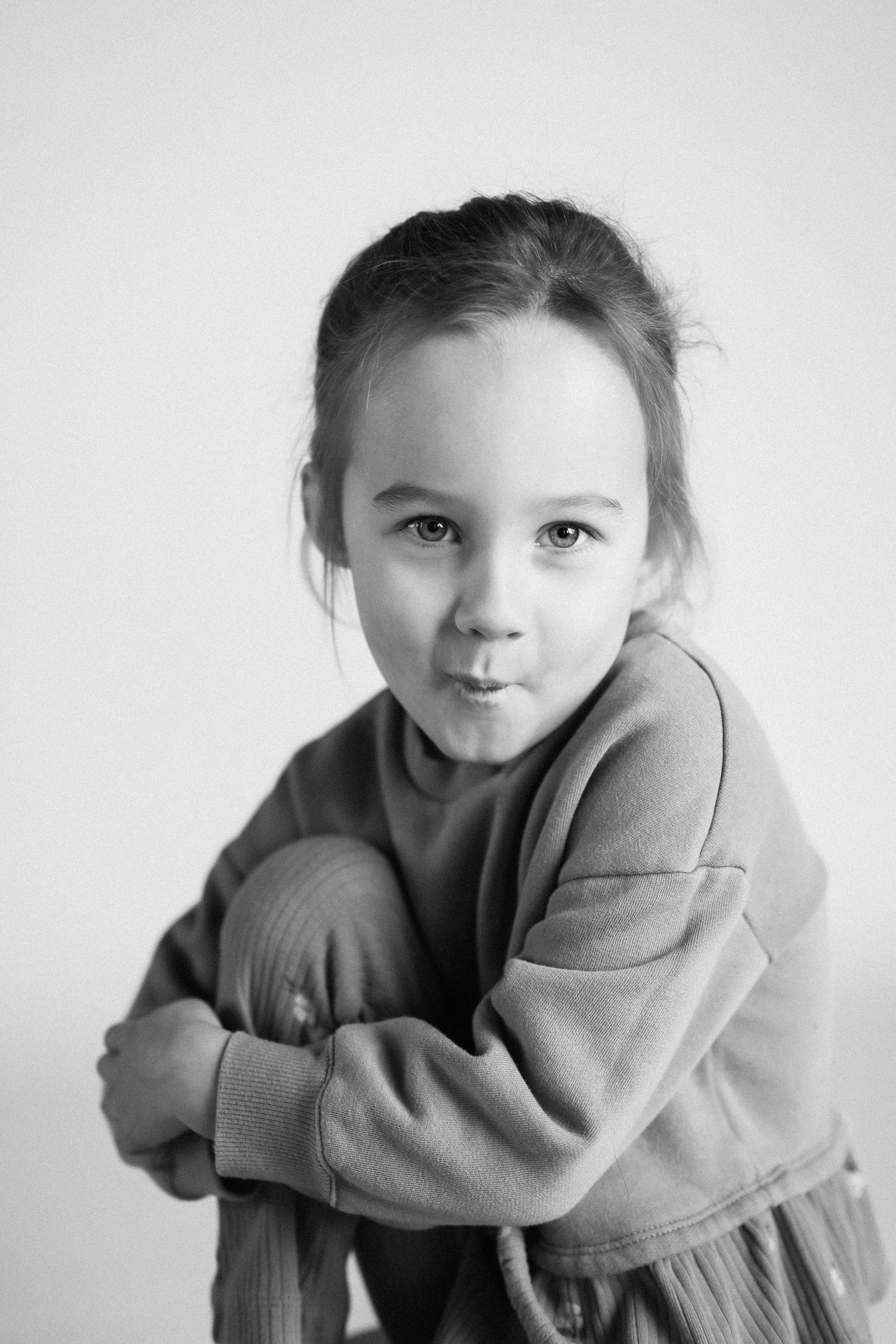 A black and white portrait of a child in a cozy sweater leaning against a white background.