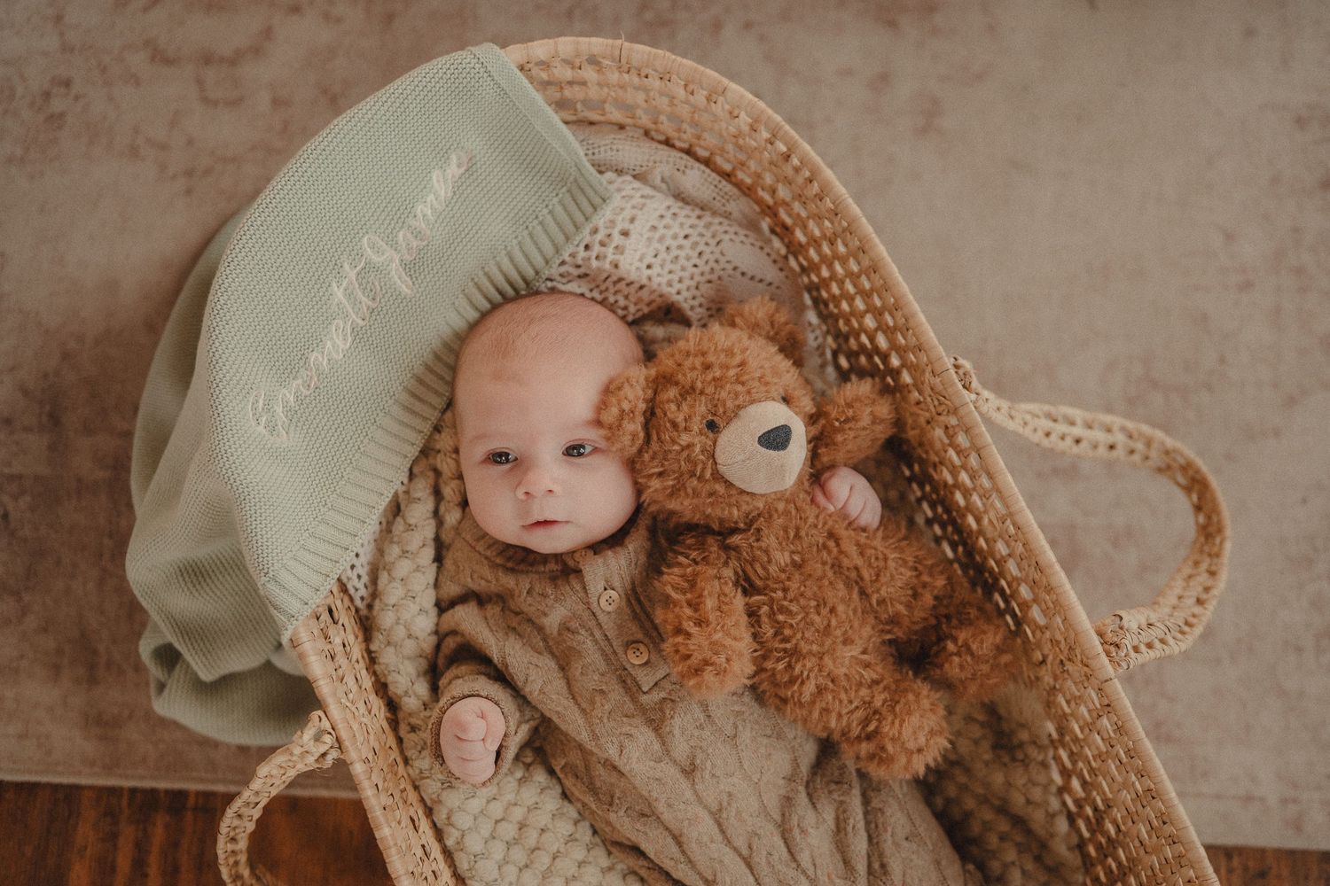 Baby lying in woven basket with soft knit blanket and teddy bear.