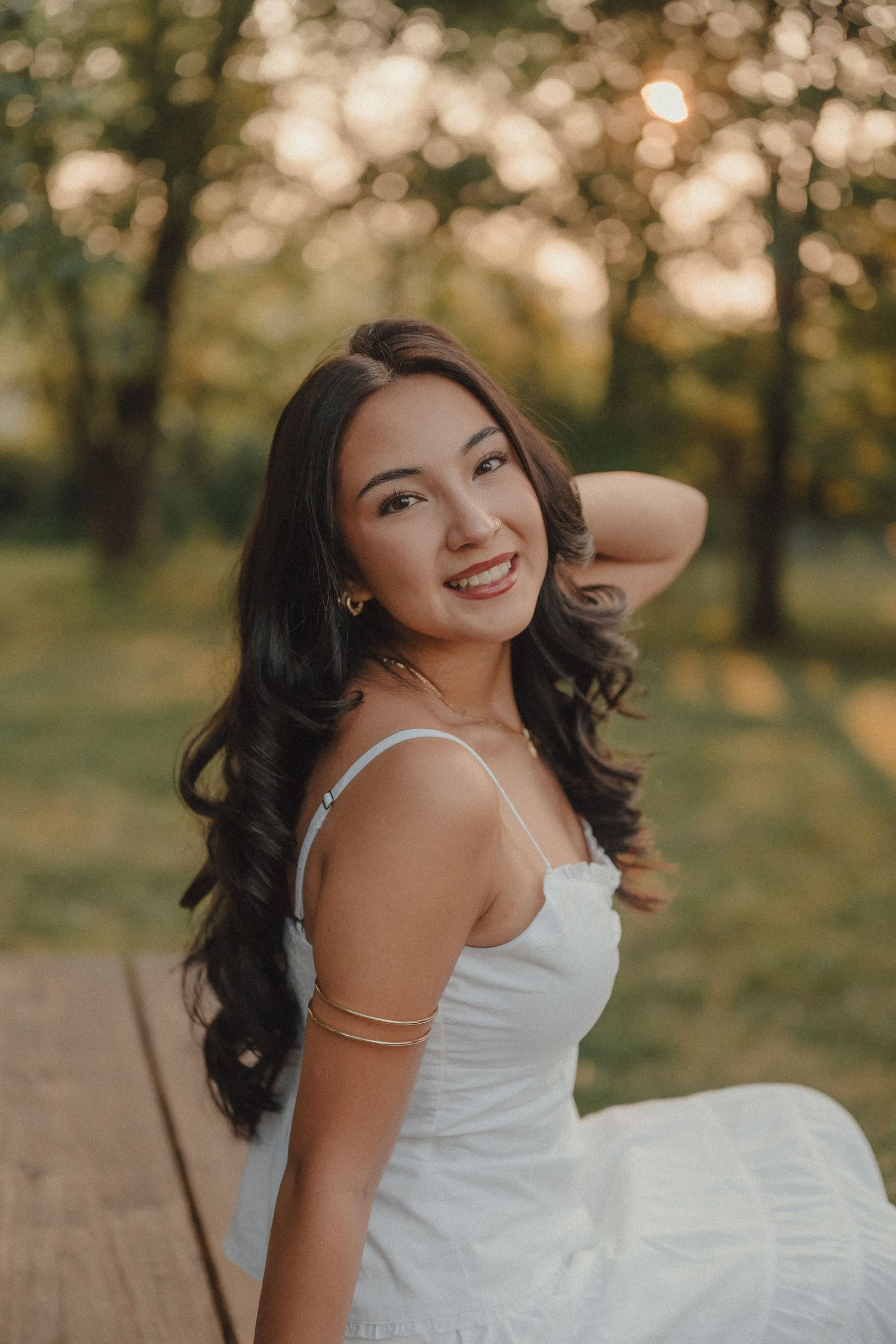 Smiling young woman with long dark wavy hair wearing a white sundress and gold arm cuff, posing outdoors at golden hour.