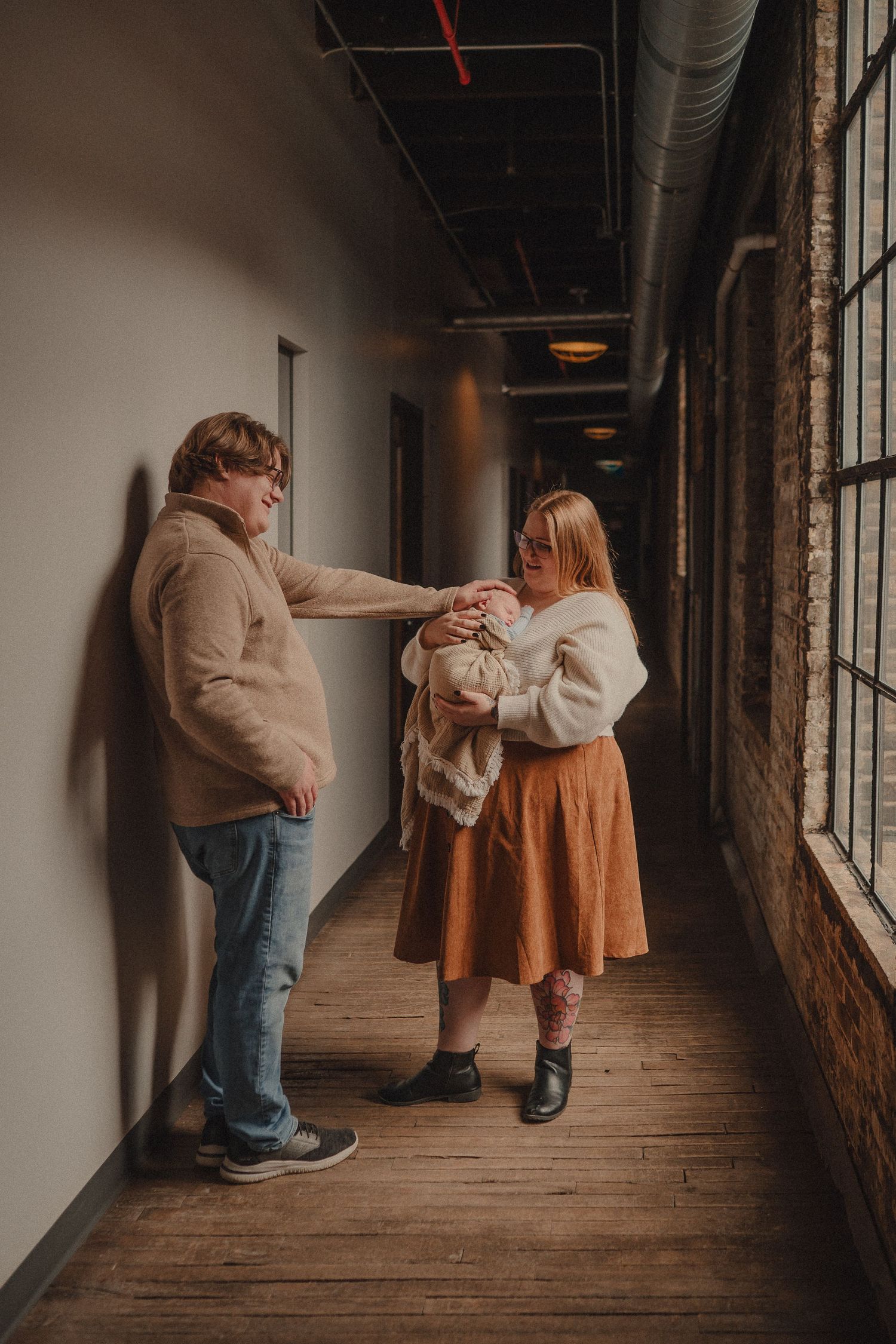 Couple in a hallway with large windows, woman in rust skirt holding newborn while partner reaches out to touch baby.