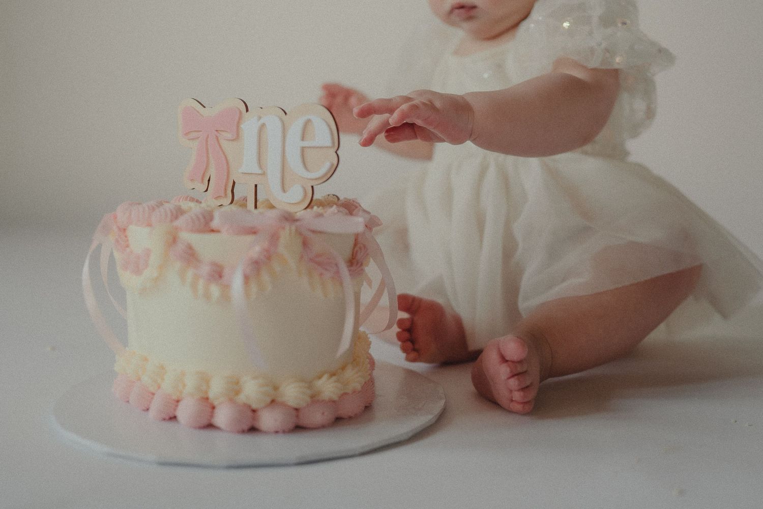 Baby girl in white dress touching her first birthday smash cake decorated in pink and gold.