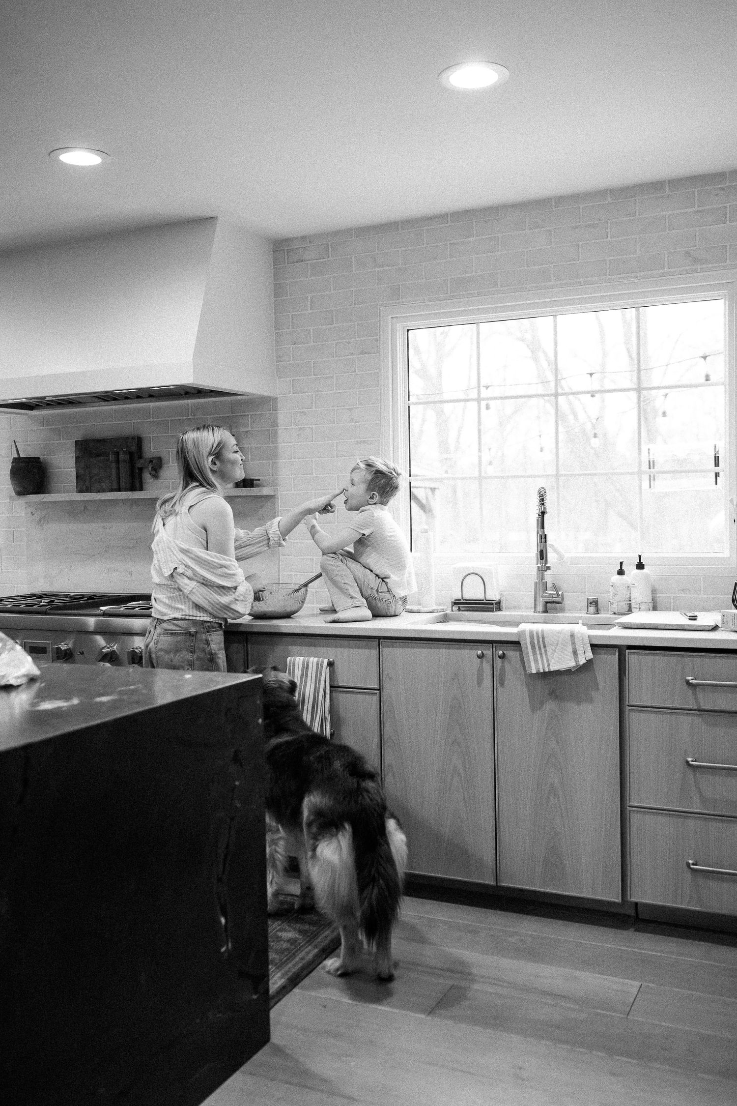 Mother and child cooking together in modern kitchen with dog nearby, black and white.