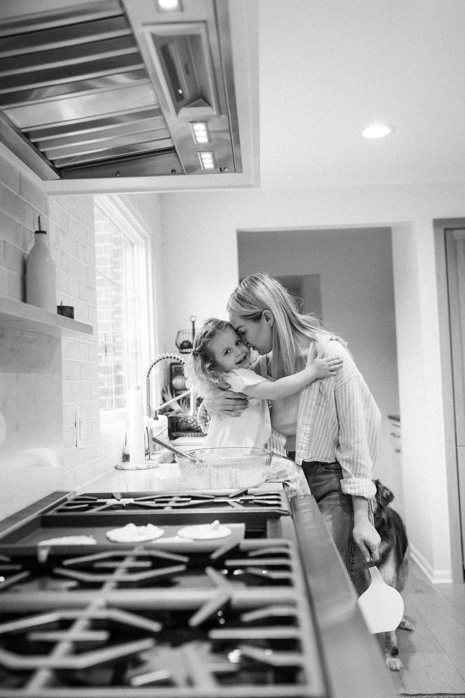 Black and white photo of a mother tenderly embracing her young daughter near a kitchen stove and sink.
