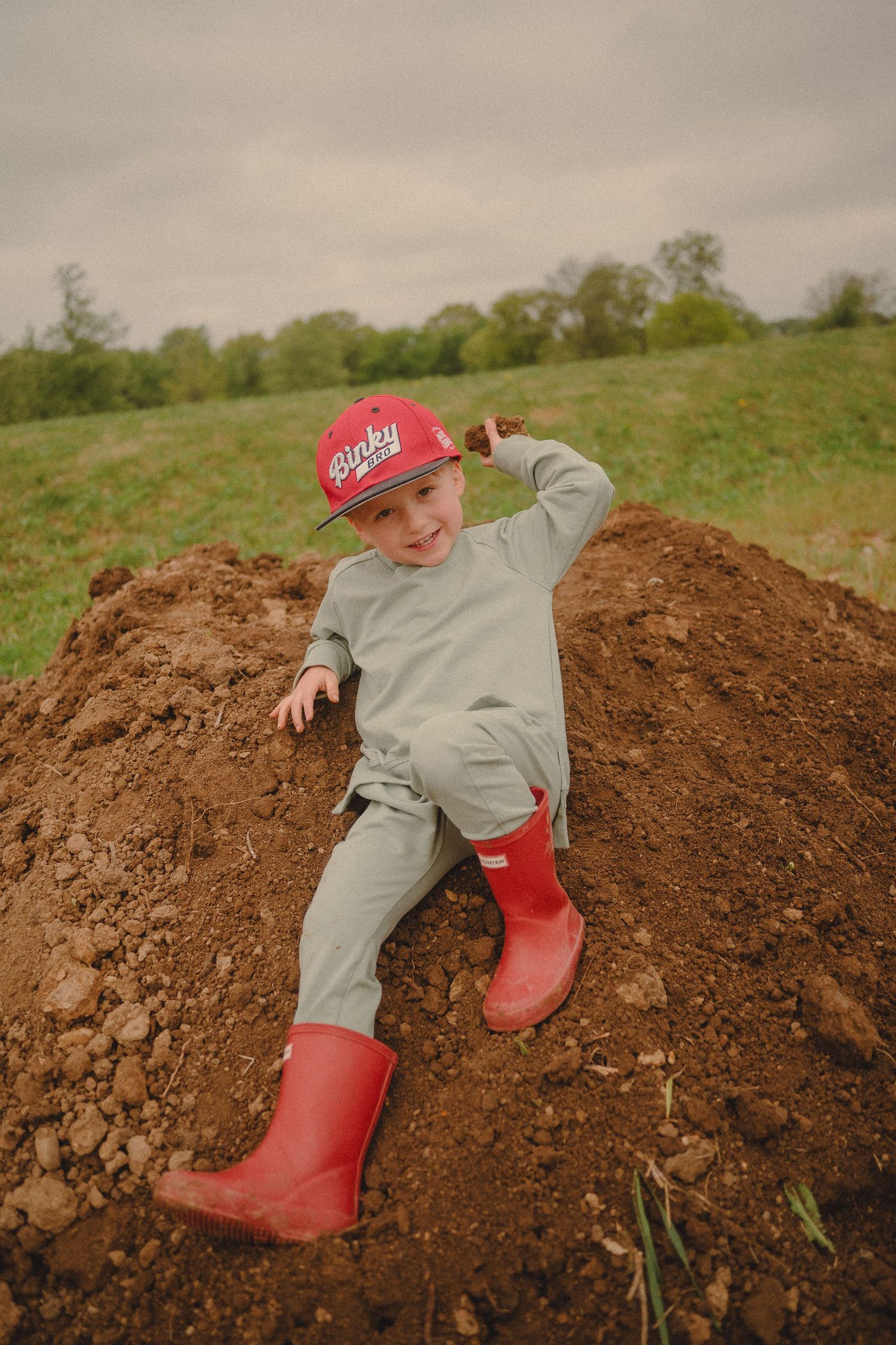 Young boy in red cap and rain boots slides down muddy dirt mound outdoors.