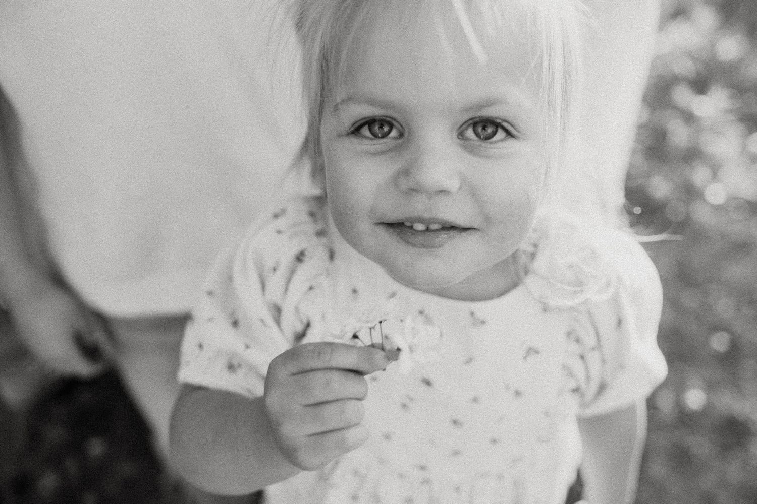 Black and white close-up portrait of a smiling blonde toddler girl holding a small flower.