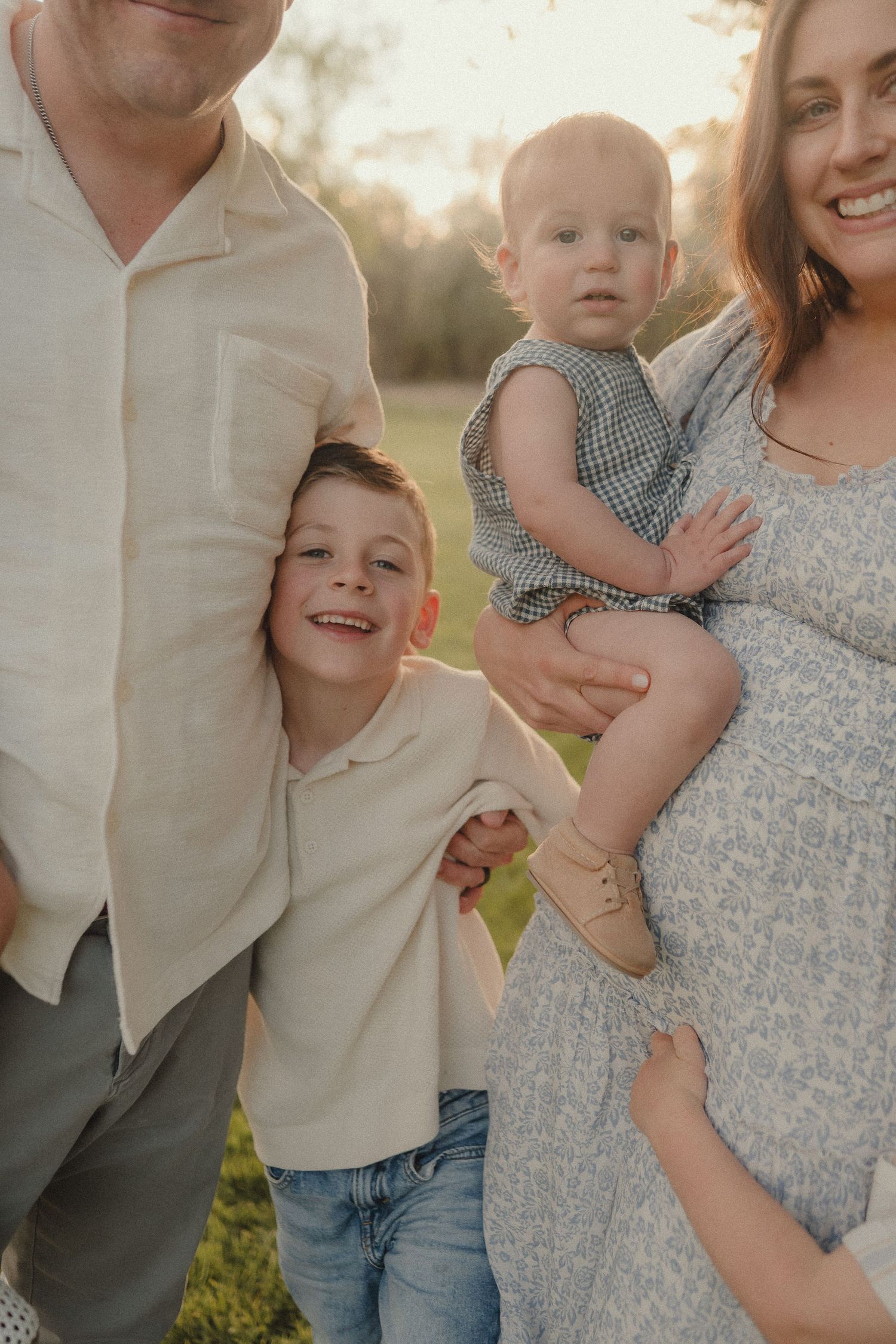 A smiling family with a pregnant mother holding a toddler and a young boy hugging his father outdoors.