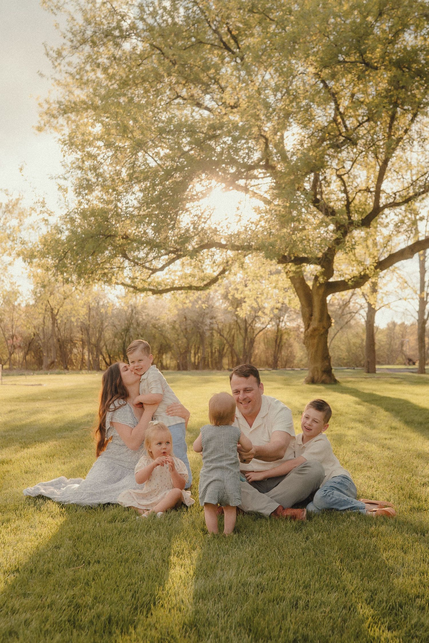 Happy family of six enjoying a sunny afternoon on the grass beneath a beautiful golden tree at sunset.