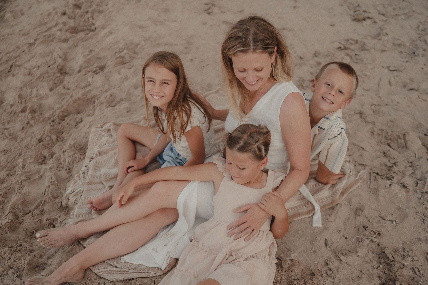 Smiling mother sits on sandy beach with three happy children on a warm sunny day.
