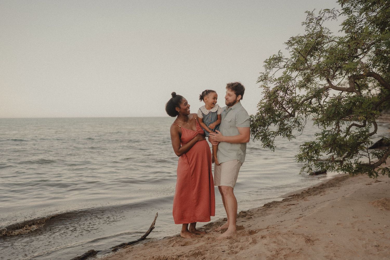 Interracial family with baby enjoying a serene beach sunset, standing near a tree on sandy shore.