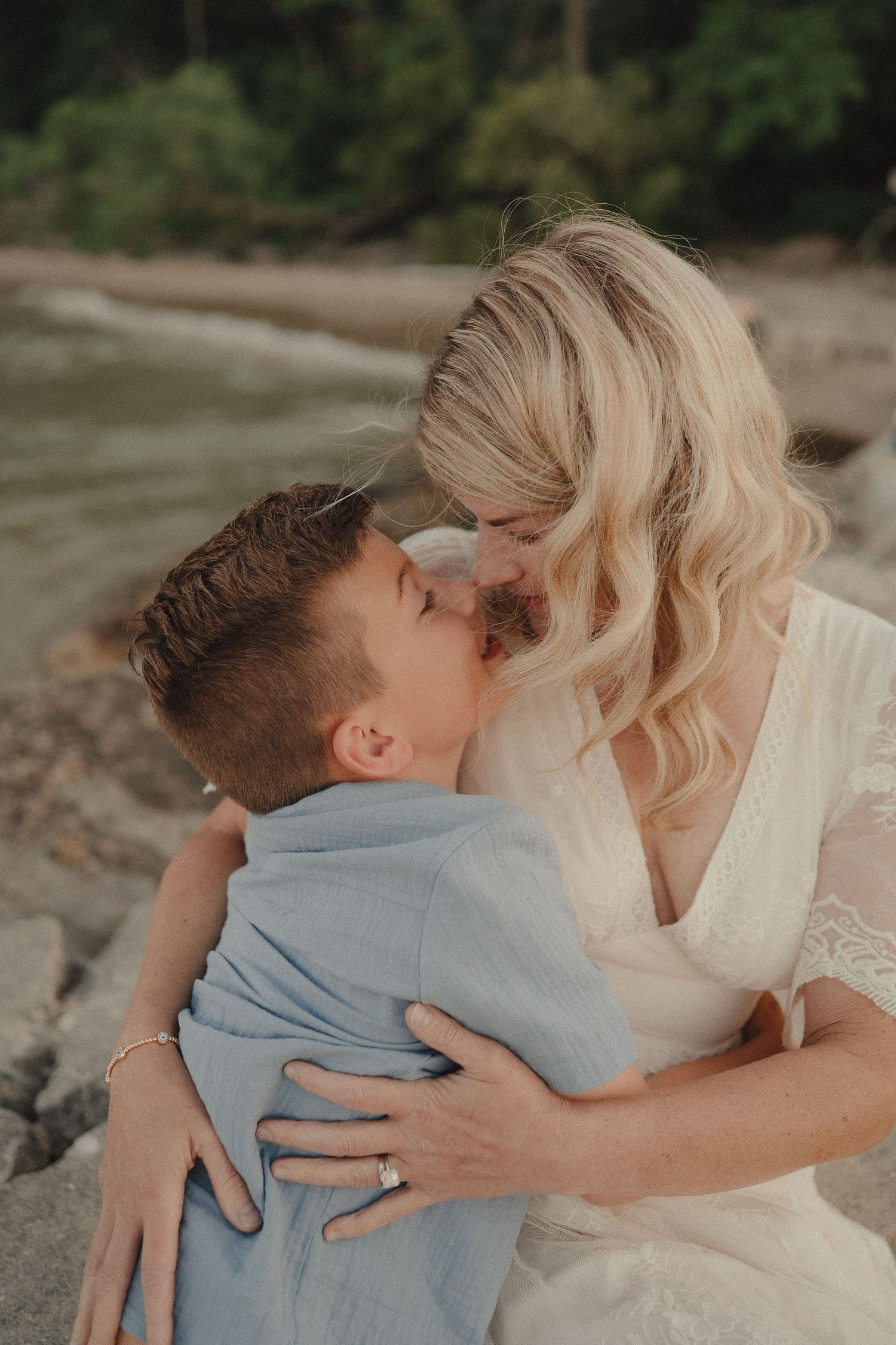 Blonde mother in white dress sharing a tender kiss with her young son on a rocky beach.