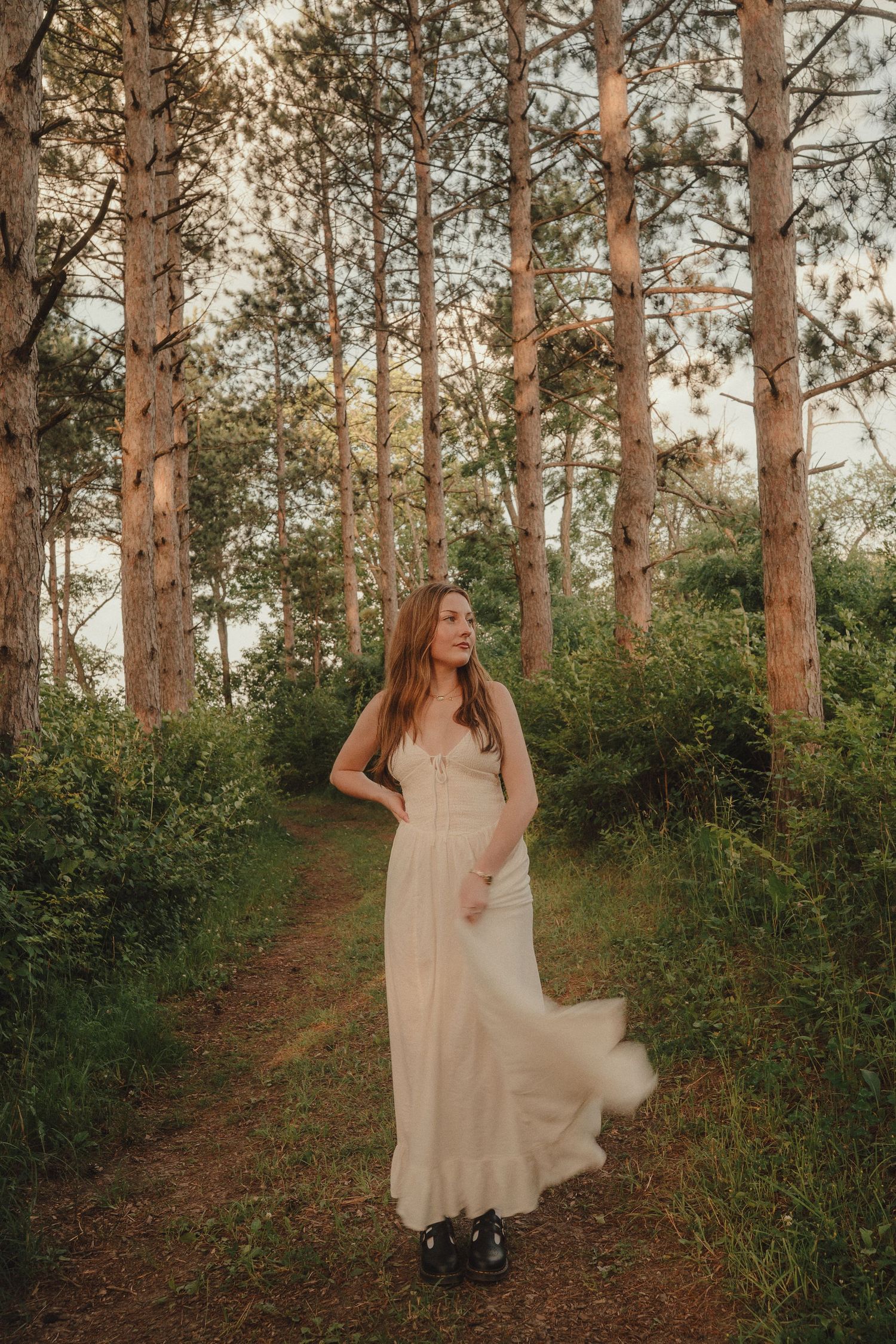 Young woman in a white maxi dress walks along a dirt path through a pine forest at golden hour.