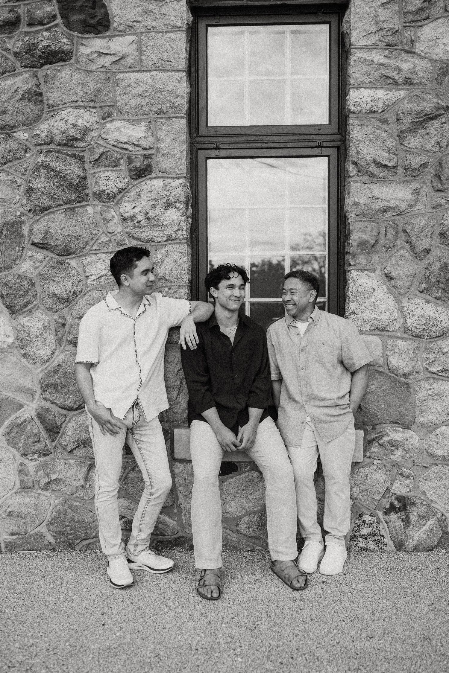 Black and white photo of three smiling men relaxing together outside a stone building with a window.
