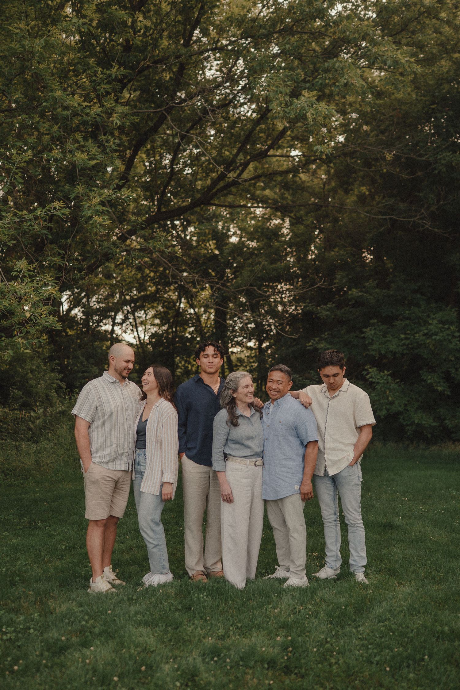 Family of six posing together on a green lawn surrounded by lush trees during golden hour.