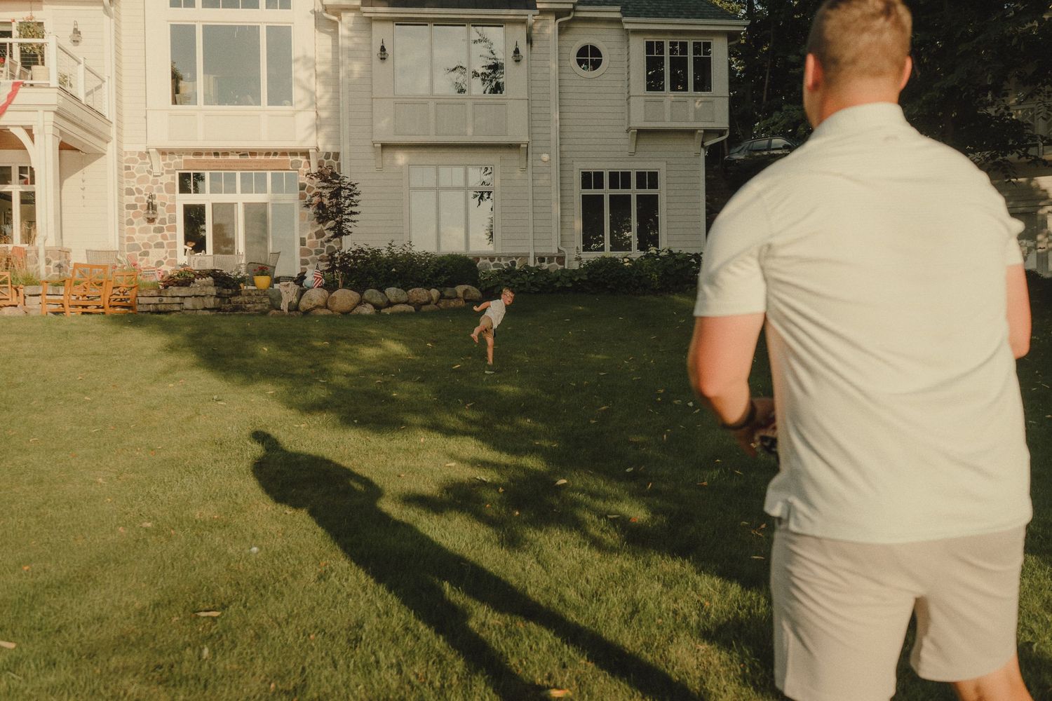 Man and child playing catch on a lush green lawn in front of a large suburban home at golden hour.