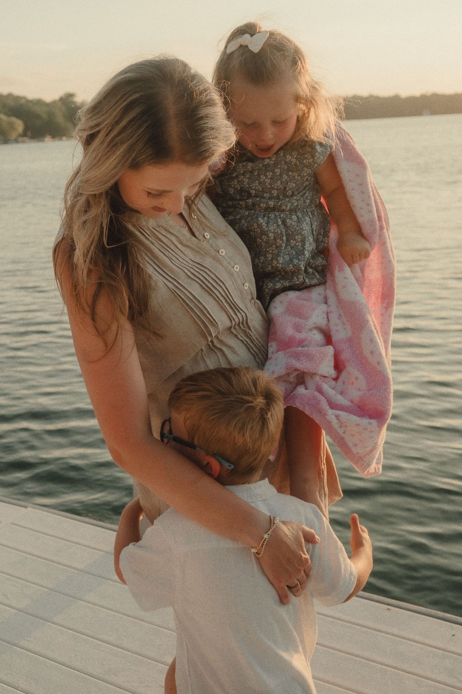 Mother holding young daughter while son hugs her waist on a lakeside dock at sunset.