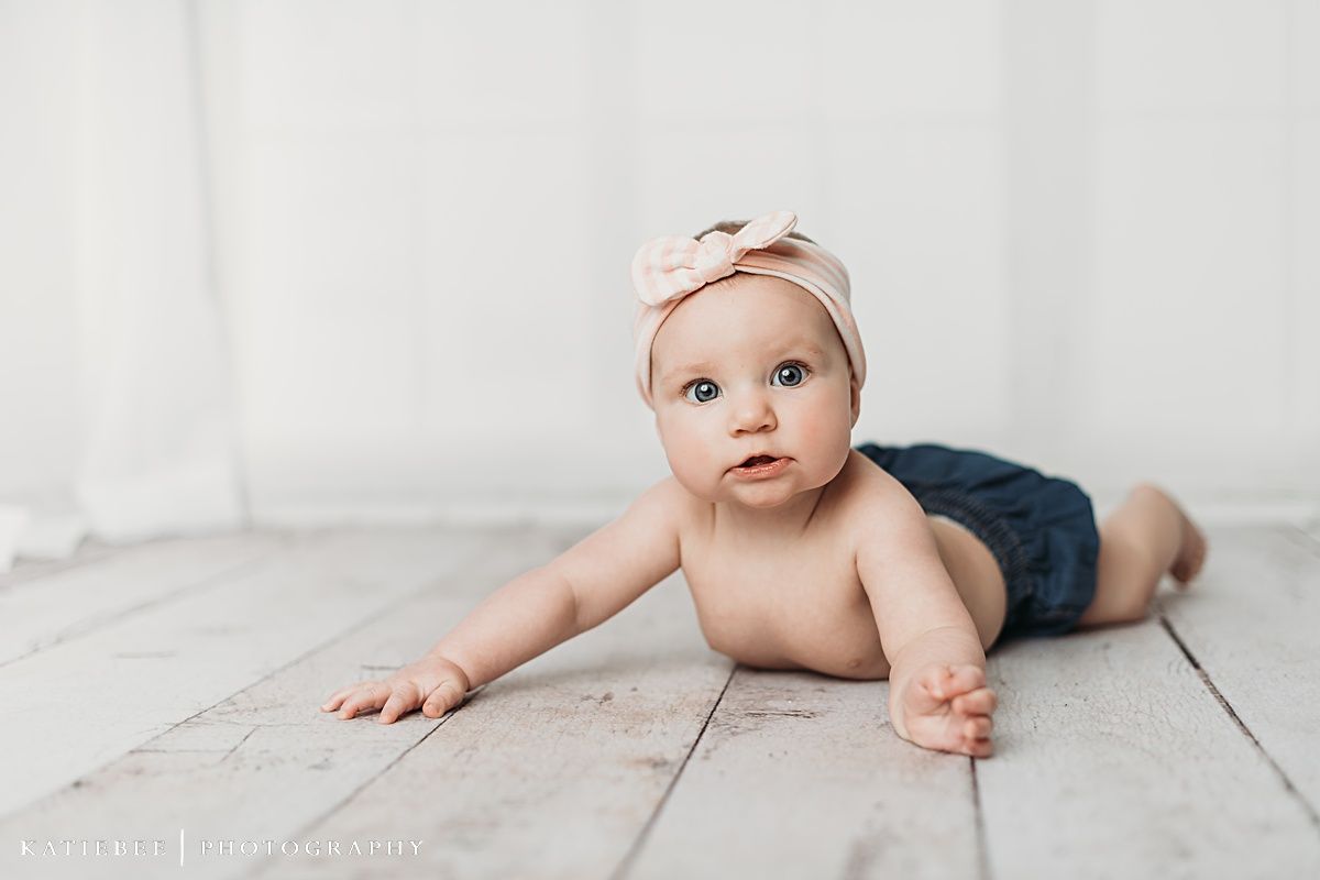 Baby wearing cream headband and dark bottoms lying on white wooden planks.