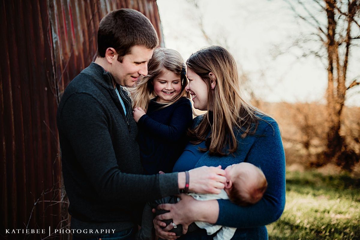 Family of four hugs close together during outdoor sunset portrait session in front of rustic barn.