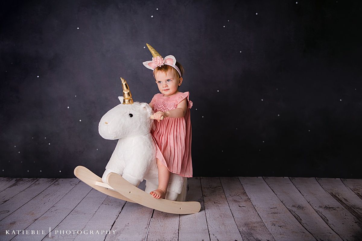 A toddler in pink plays on a white plush rocking unicorn against a dark studio backdrop.
