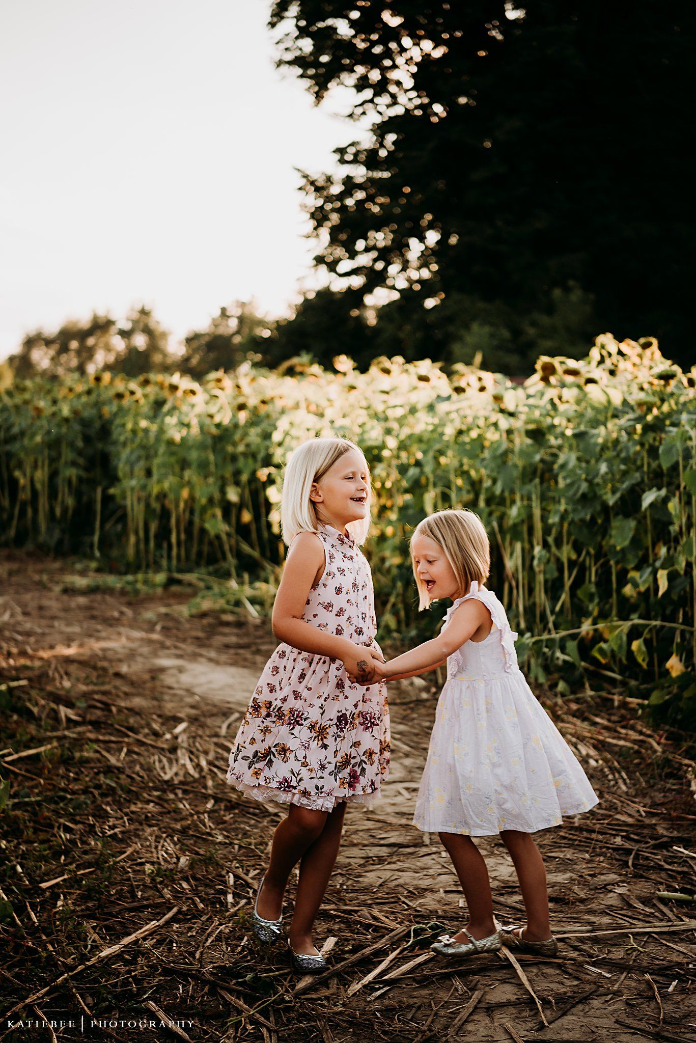 Two children in summer dresses playing together on a path beside towering sunflowers at sunset.