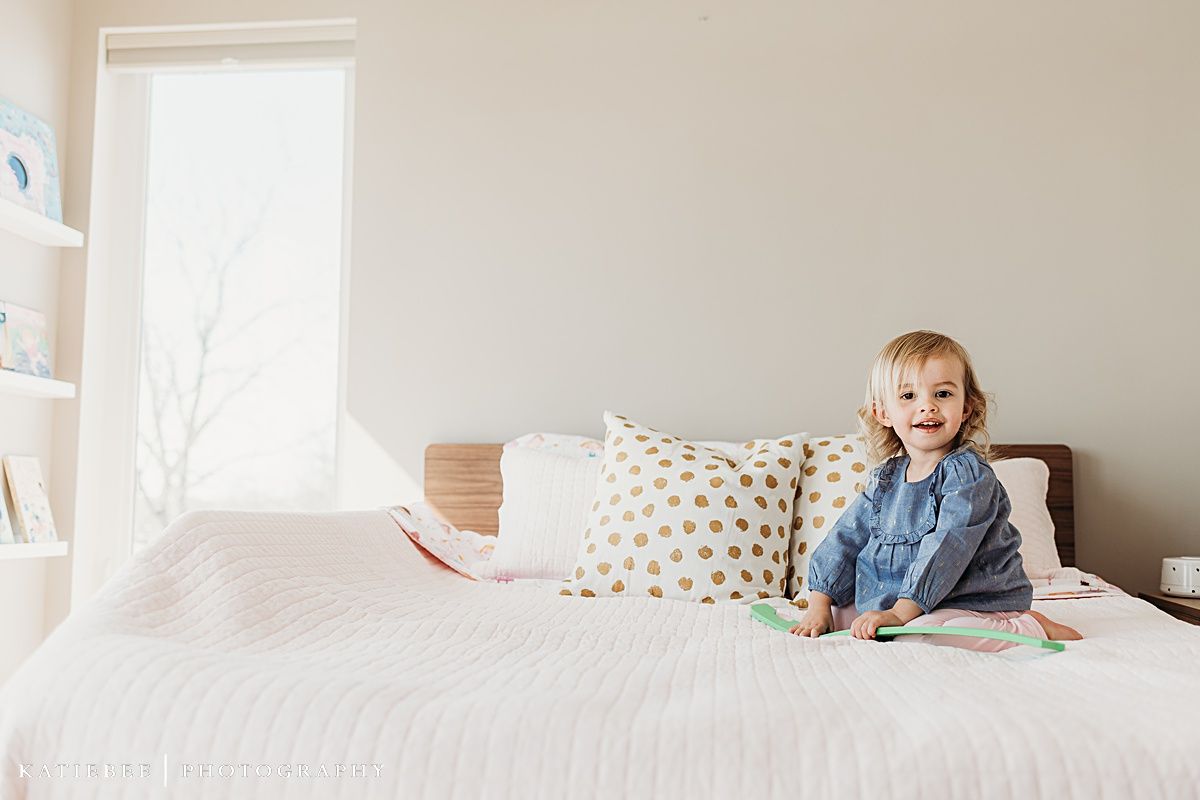 Small child in blue dress sits happily on white bed with decorative pillows.