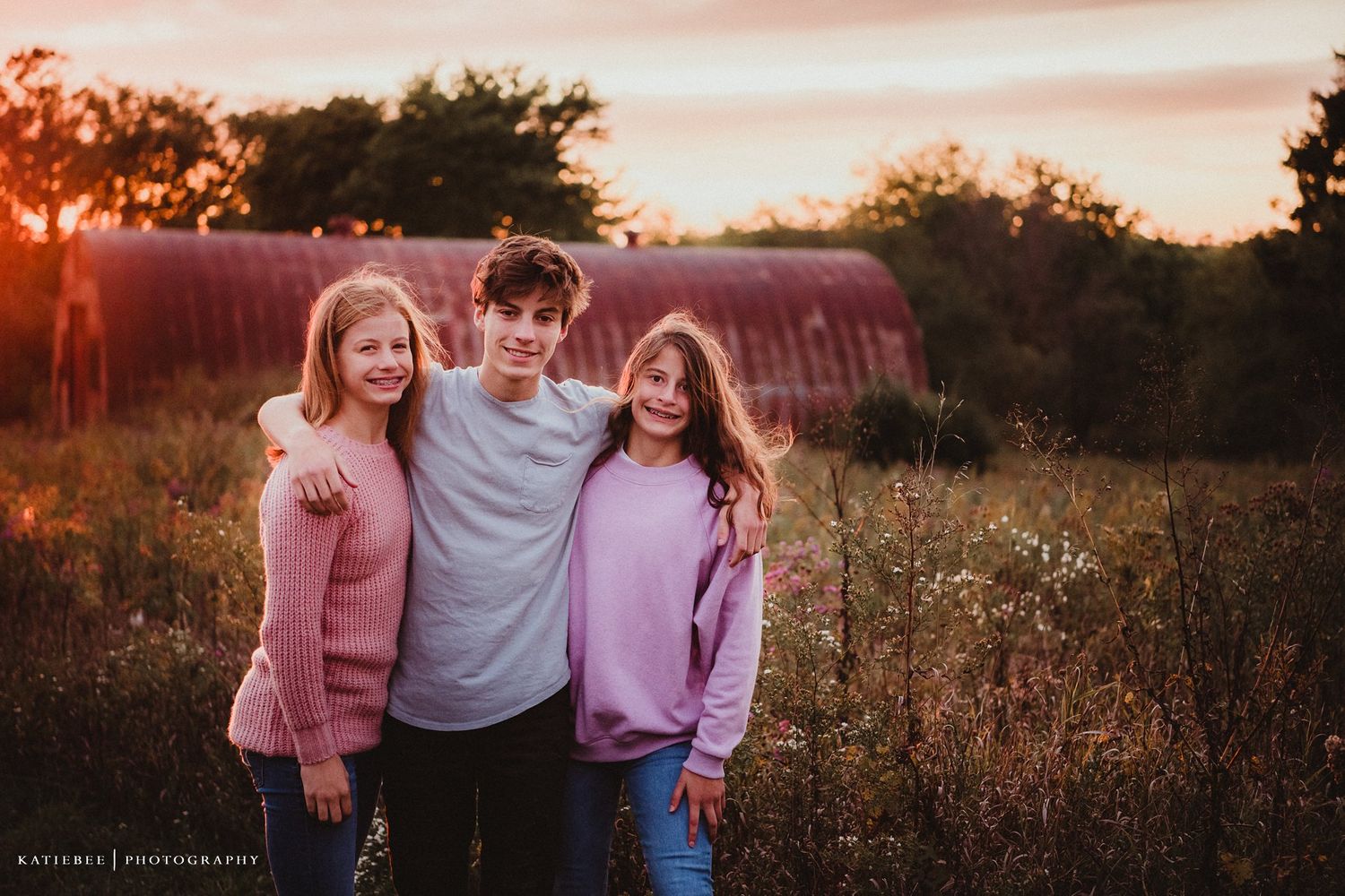 Family portrait at sunset with pink sky and red barn in background.