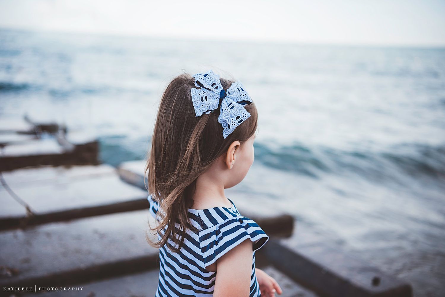 A child in striped dress with bow looks out at the ocean from a rocky shoreline.