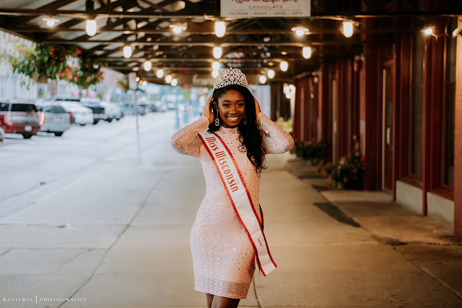 Person wearing pink dress and sash poses under evening city lighting.