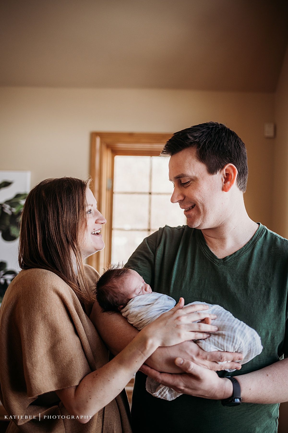 Parents cradle newborn baby while sharing intimate moment near window.