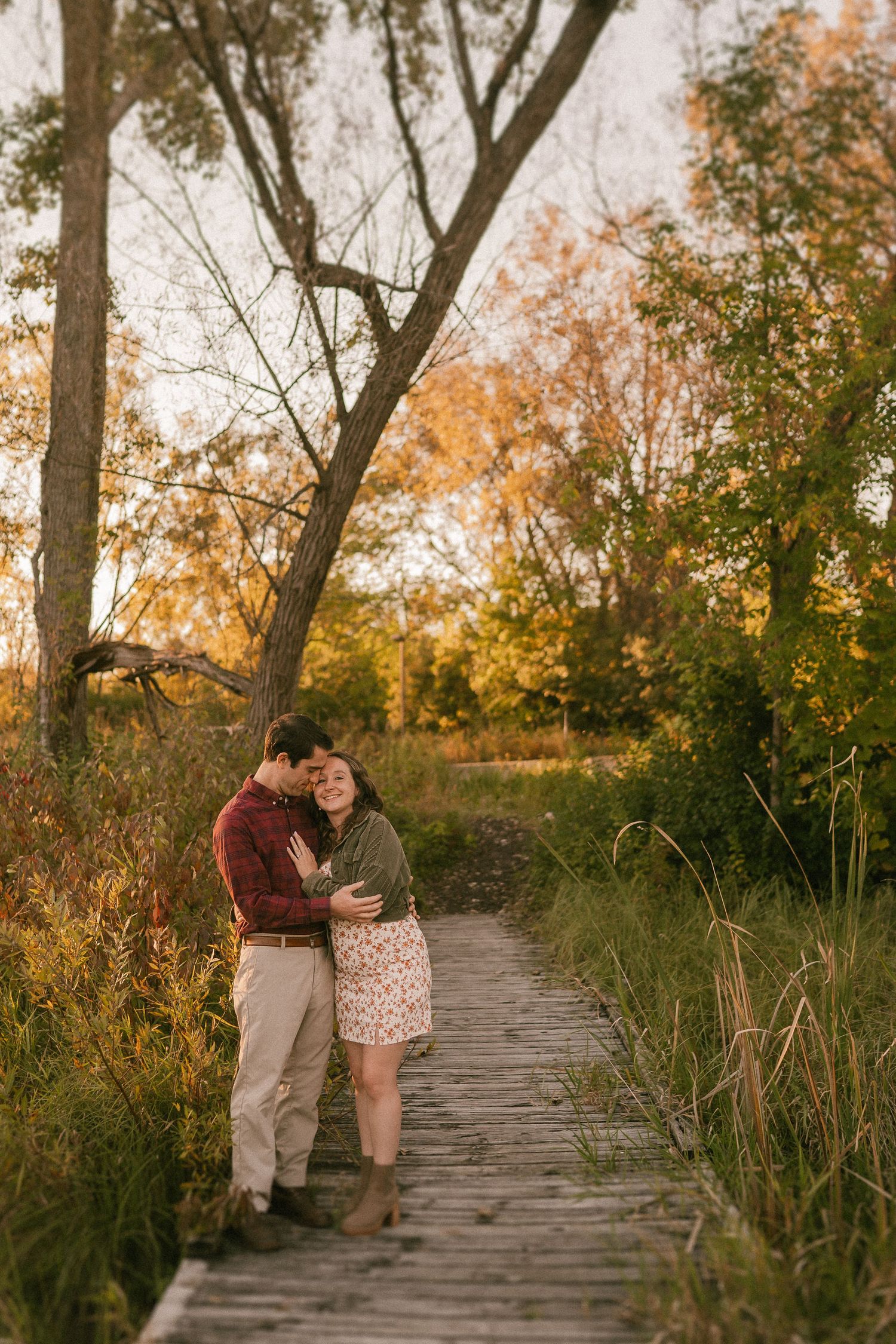 A tender moment captured on a wooden boardwalk during sunset with autumn trees in the background.