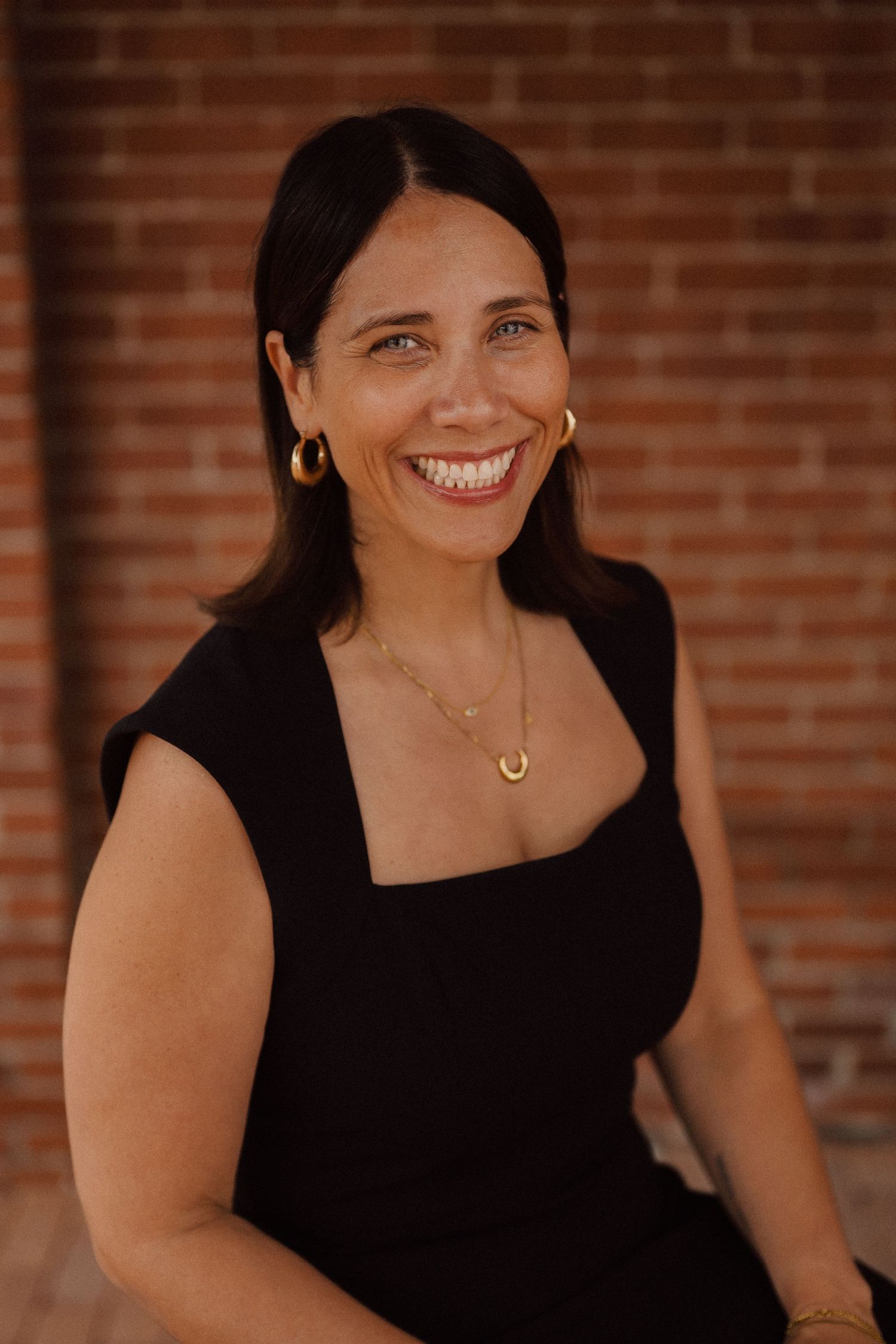 Professional headshot of someone in a black sleeveless top smiling against a brick wall.