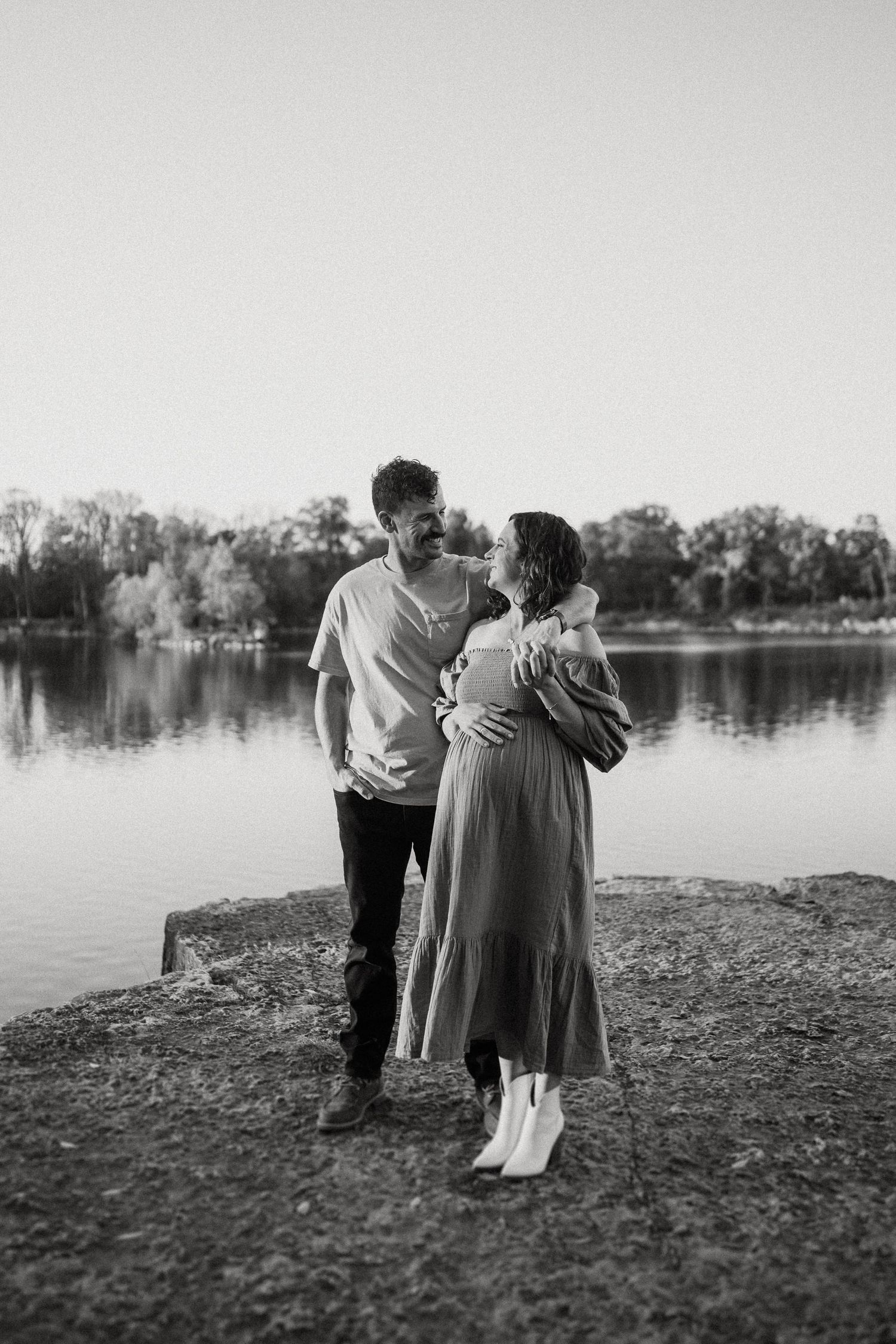 Black and white portrait of expecting couple embracing on lakeside shore.