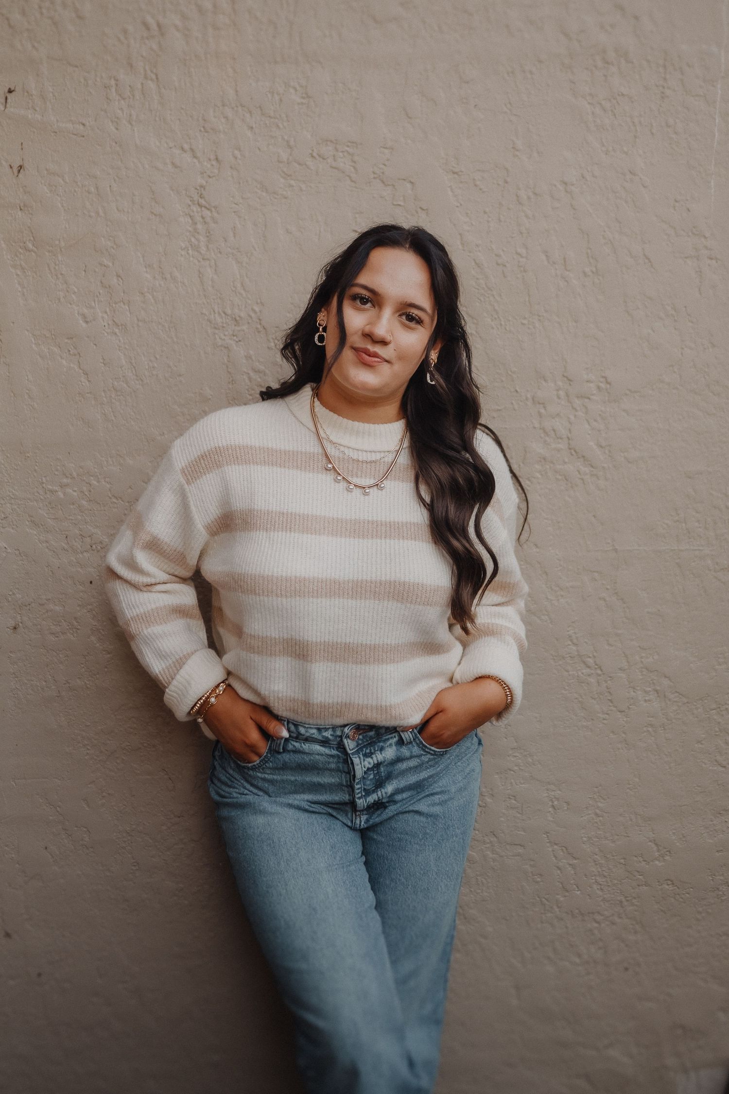 Woman in striped cream sweater and blue jeans poses against textured beige wall with a subtle smile.