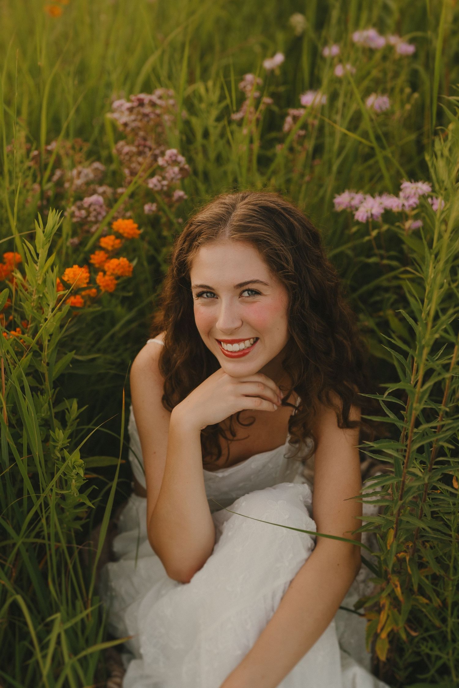 A woman in a white dress sits among vibrant wildflowers and tall grass during a summer photoshoot.