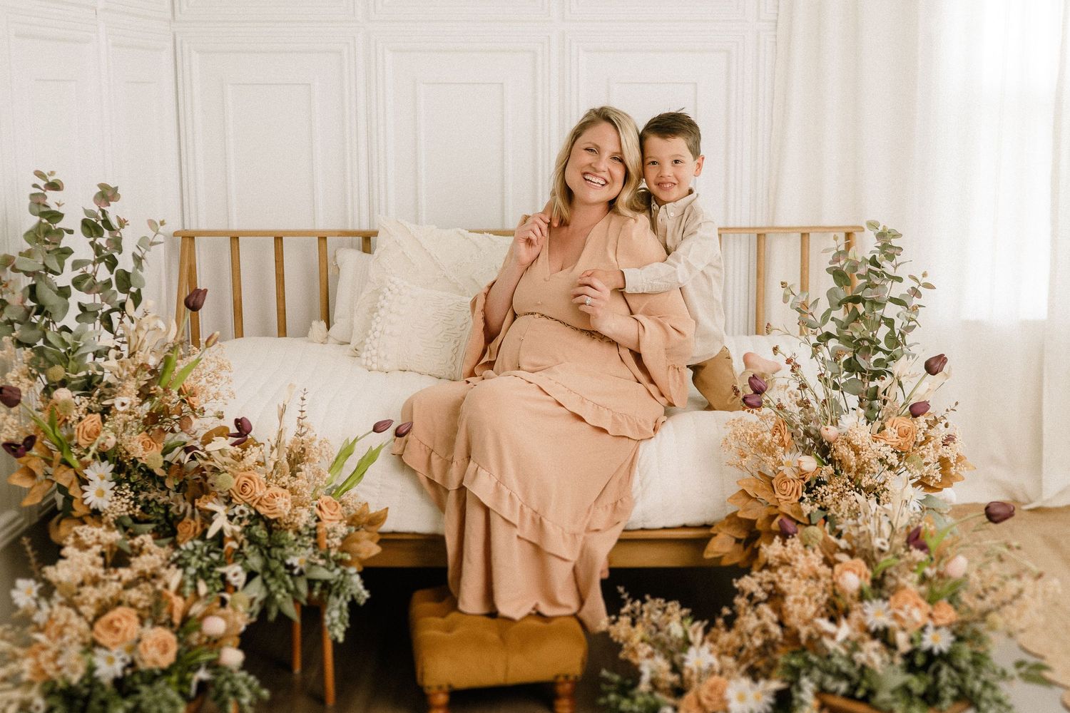 Beautiful floral arrangement surrounds a mother and child sitting on a bed in a light and airy room with neutral decor.