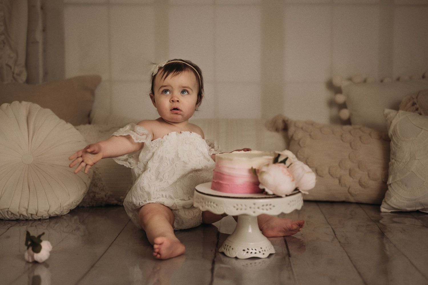 A toddler in white dress reaching toward pink cake on ornate white pedestal stand.