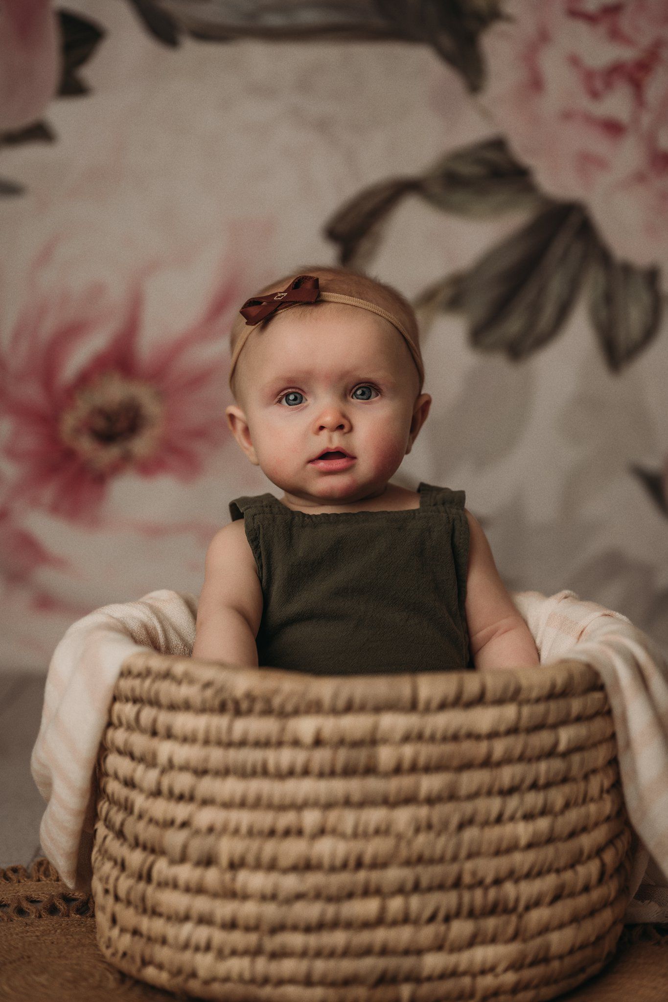 Baby wearing dark tank top sits in basket against floral background.