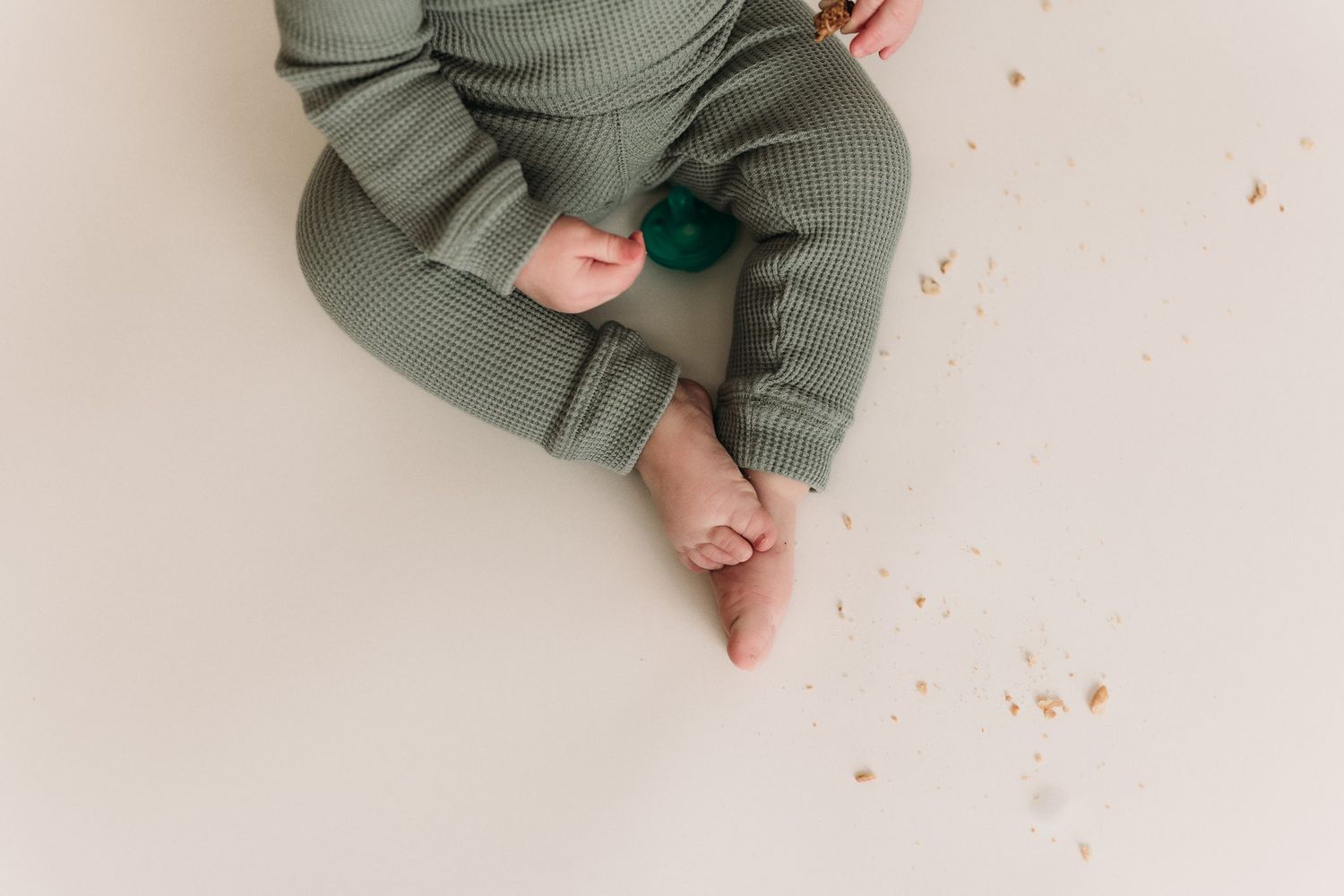 Baby in sage green outfit seated on white background looking at camera.
