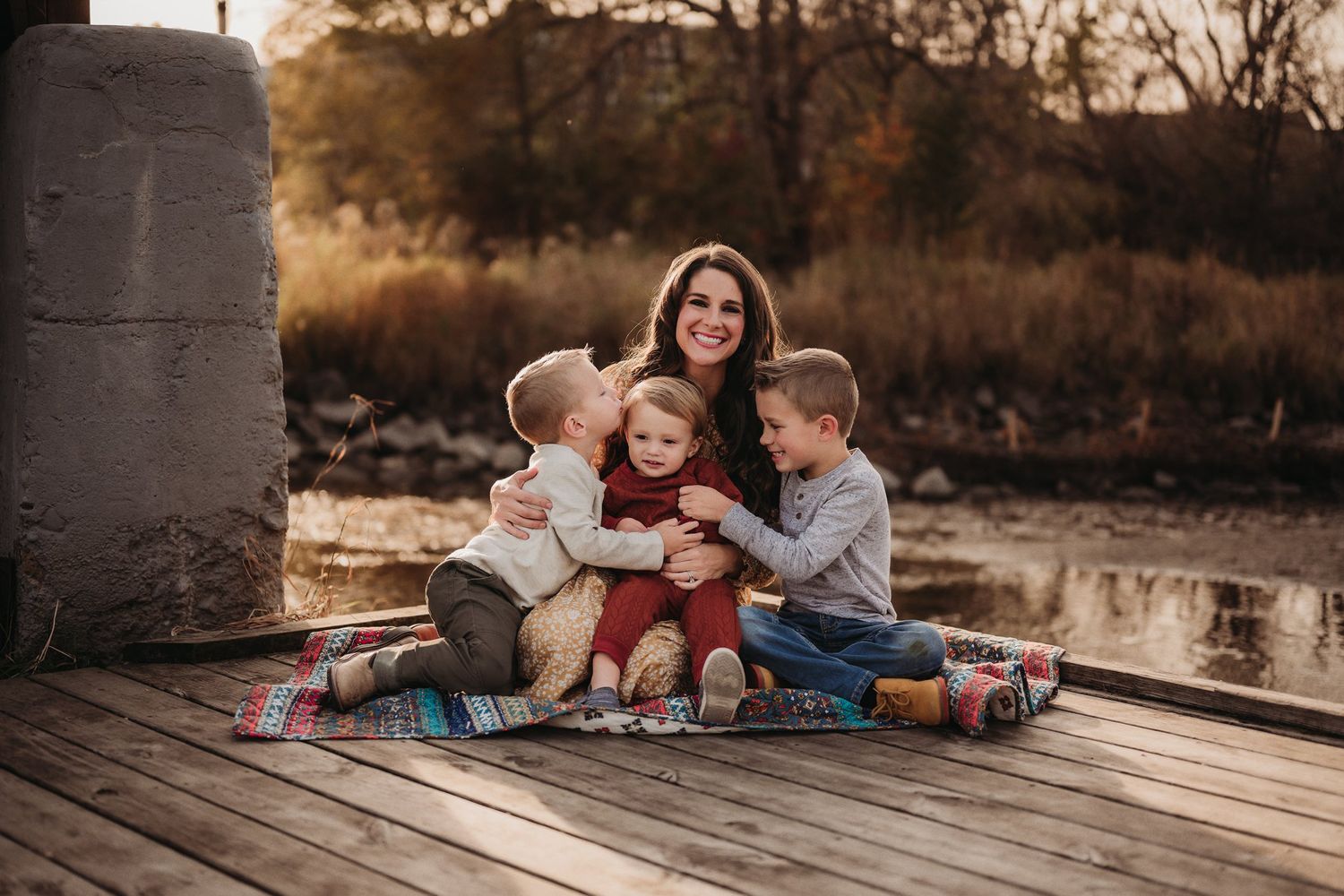 A family sits together on a blanket on a wooden dock at sunset.