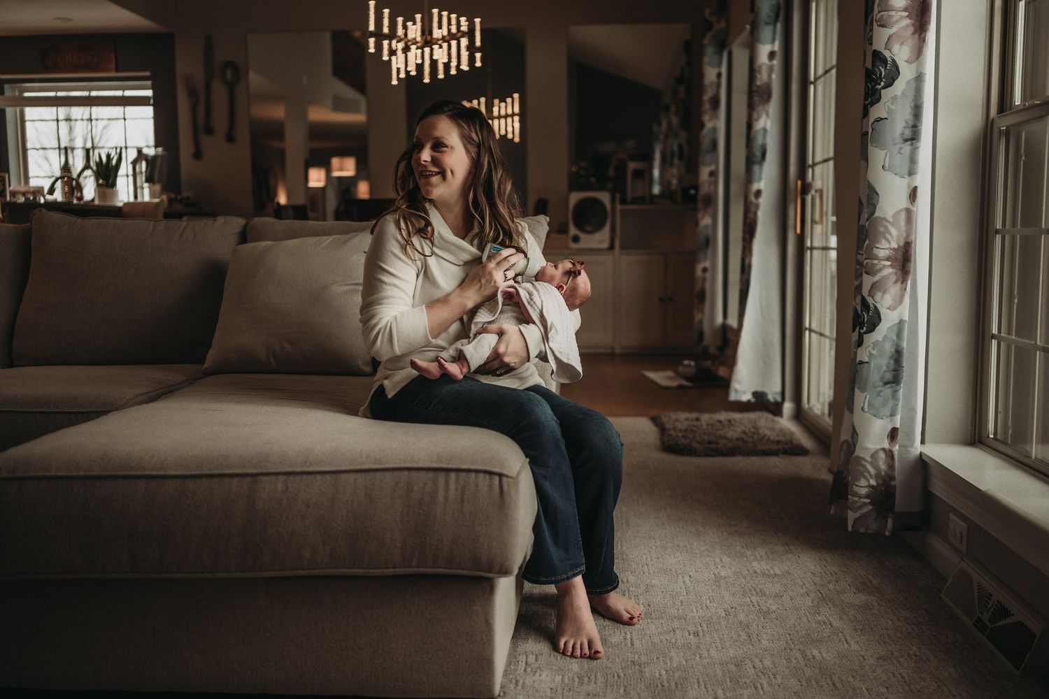 Warm lifestyle photo of parent and baby relaxing on beige couch by window.