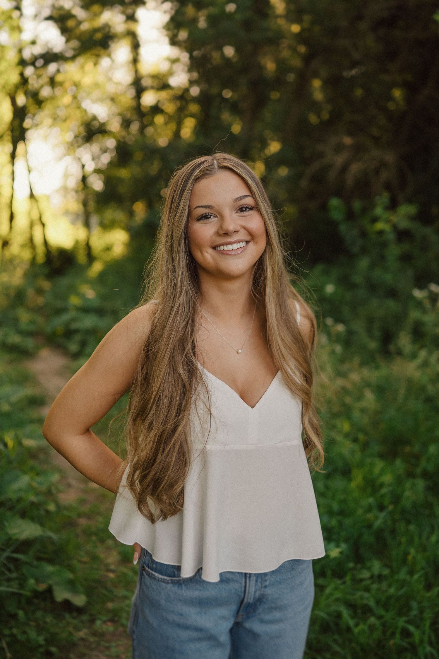Natural outdoor portrait in white camisole and jeans with sunlit forest background.