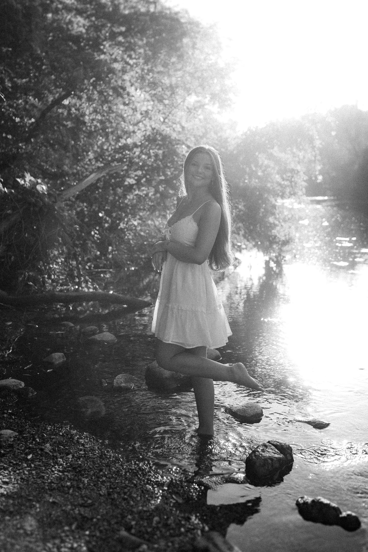 Black and white photo of a figure standing on rocks by a tranquil stream.
